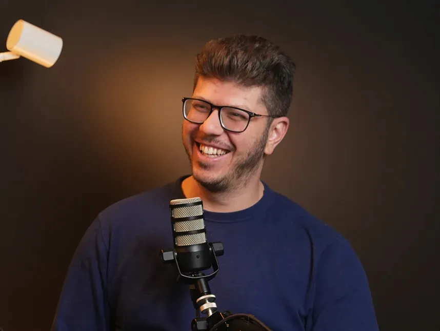 Charles Homsi qui rit, lunettes et chandail marine, derrière un micro de balado dans son studio.