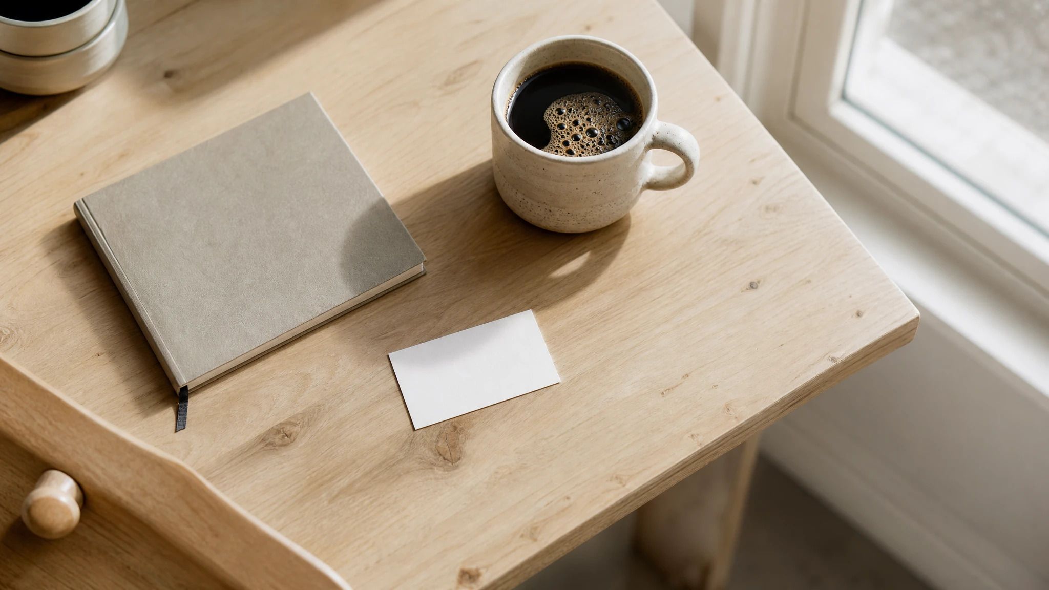 Pale wooden desk by a window, with a notebook, a cup of coffee and a blank business card.